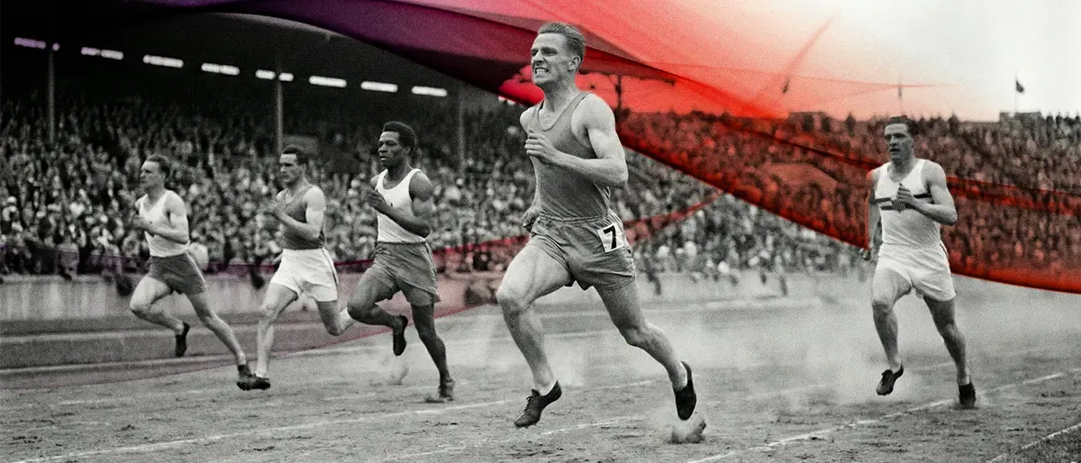 A black and white vintage-style photograph of several male runners sprinting on a dirt track in front of a crowded stadium.